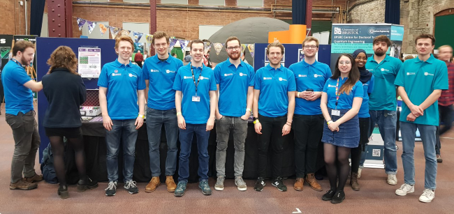 一群量子工程专业的学生在展台上穿着相配的t恤，微笑着摆姿势对着相机。 